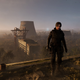 A man stands at the edge of a decrepit building. A vast expanse of wilderness looms behind him, interspersed with concrete silos.